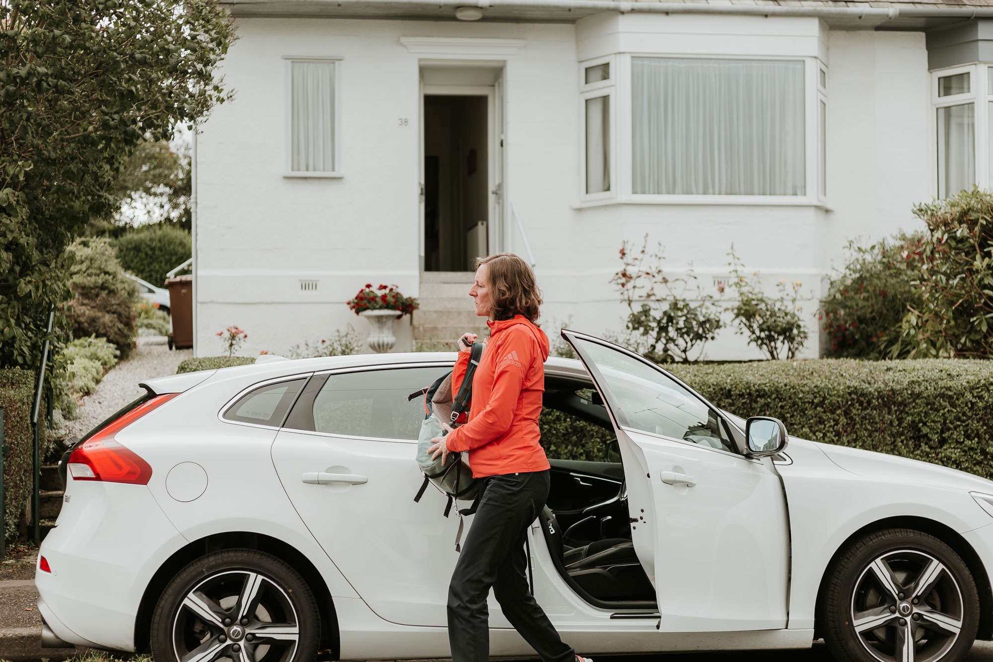 Physiotherapist leaving a car and putting on a rucksack before attending a home physiotherapy assessment appointment outside a white house.